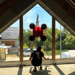 Montaż okna o nietypowym kształcie w surowym stanie poddasza, widoczny pracownik w trakcie instalacji, na zewnątrz dźwig z przyssawkami do szkła i zabudowa domów jednorodzinnych.