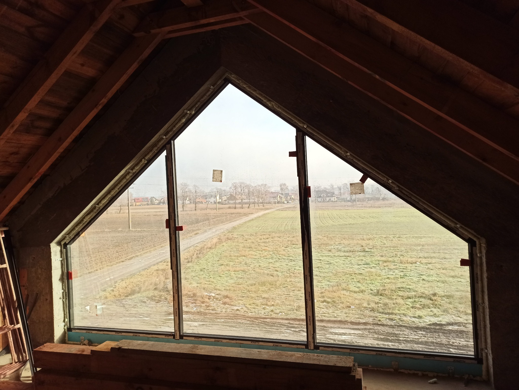 Trójkątne okno aluminiowe w surowym stanie poddasza, widok na pole i zabudowania, montaż w trakcie prac wykończeniowych.