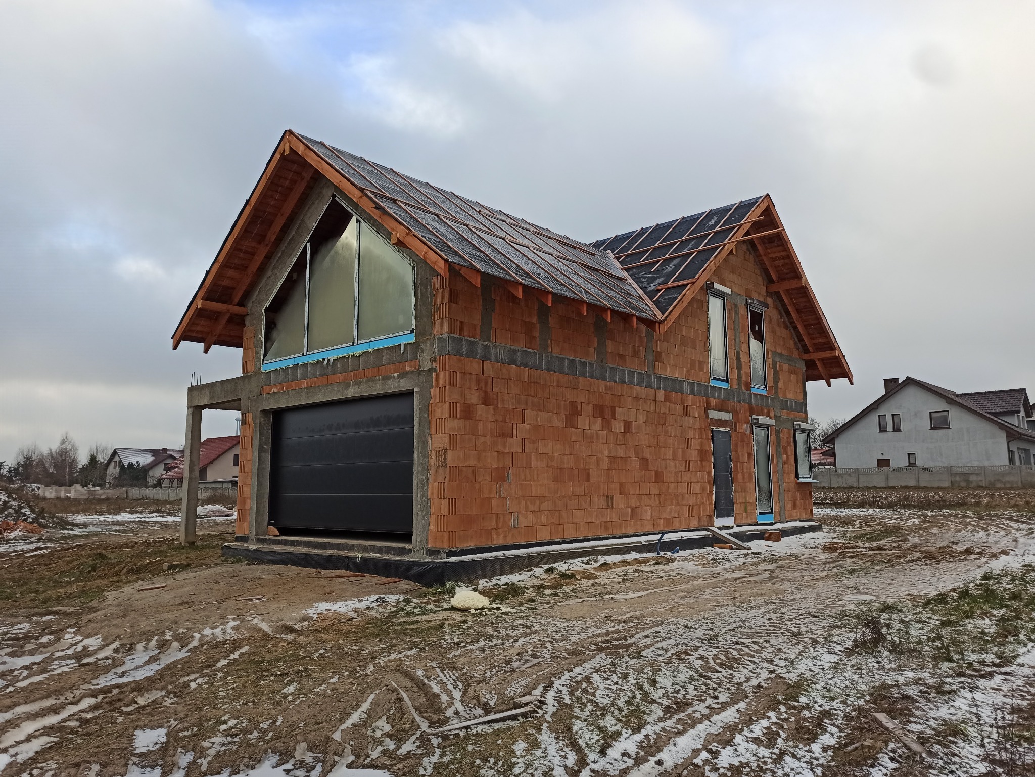 Surowy stan domu jednorodzinnego w trakcie budowy, widoczna czarna brama garażowa, niezadaszony dach z częściowo ułożoną dachówką, okna zabezpieczone folią, oraz pobrudzony śniegiem teren budowy.