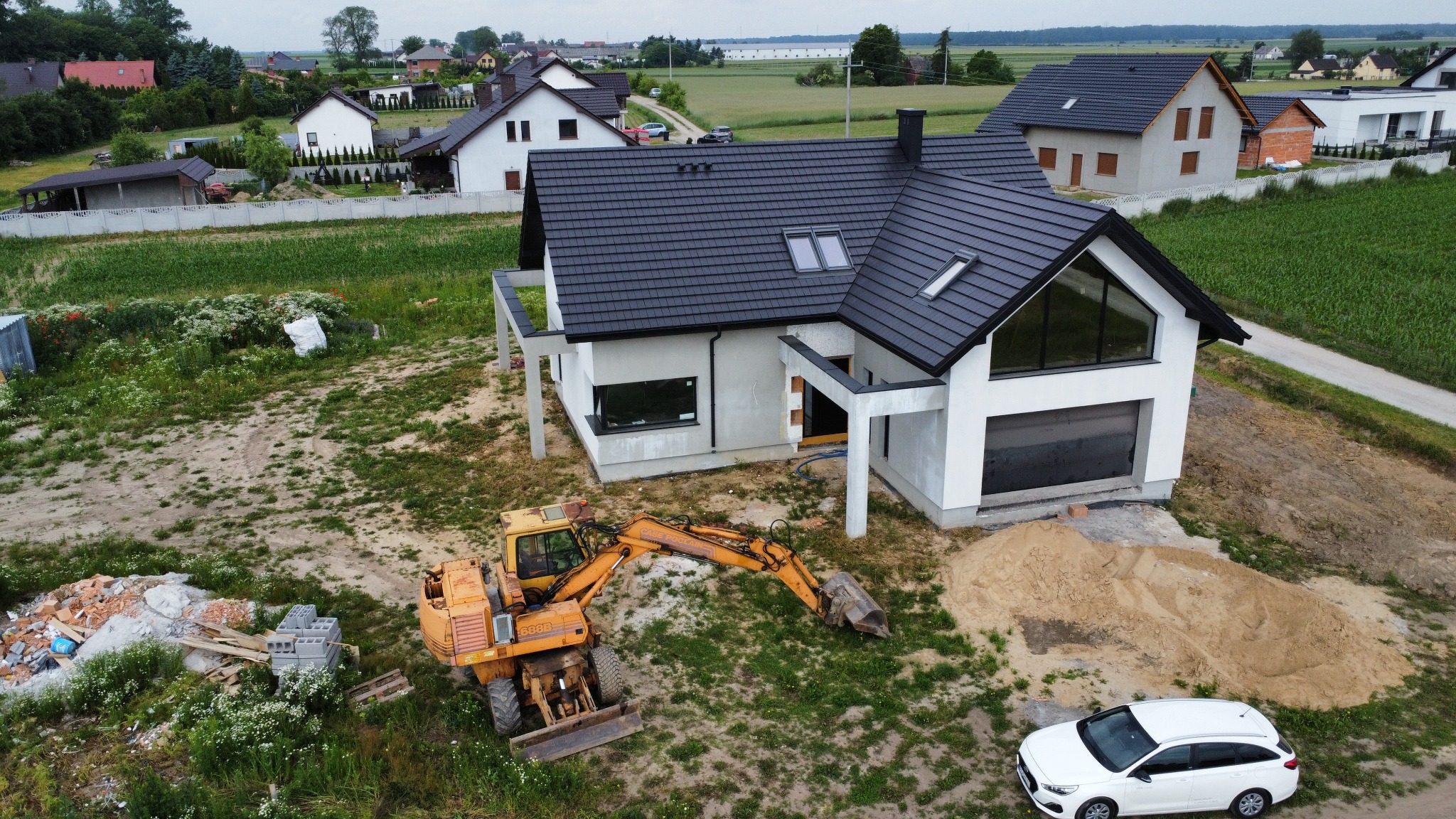 Nowoczesny dom jednorodzinny w trakcie budowy, widok z góry, z widocznym żółtym koparko-ładowarką i stertą piasku na posesji.