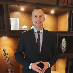 Portret uśmiechniętego mężczyzny w garniturze, stojącego na tle drewnianej ściany z podświetlanymi półkami, na których znajdują się dekoracyjne przedmioty.