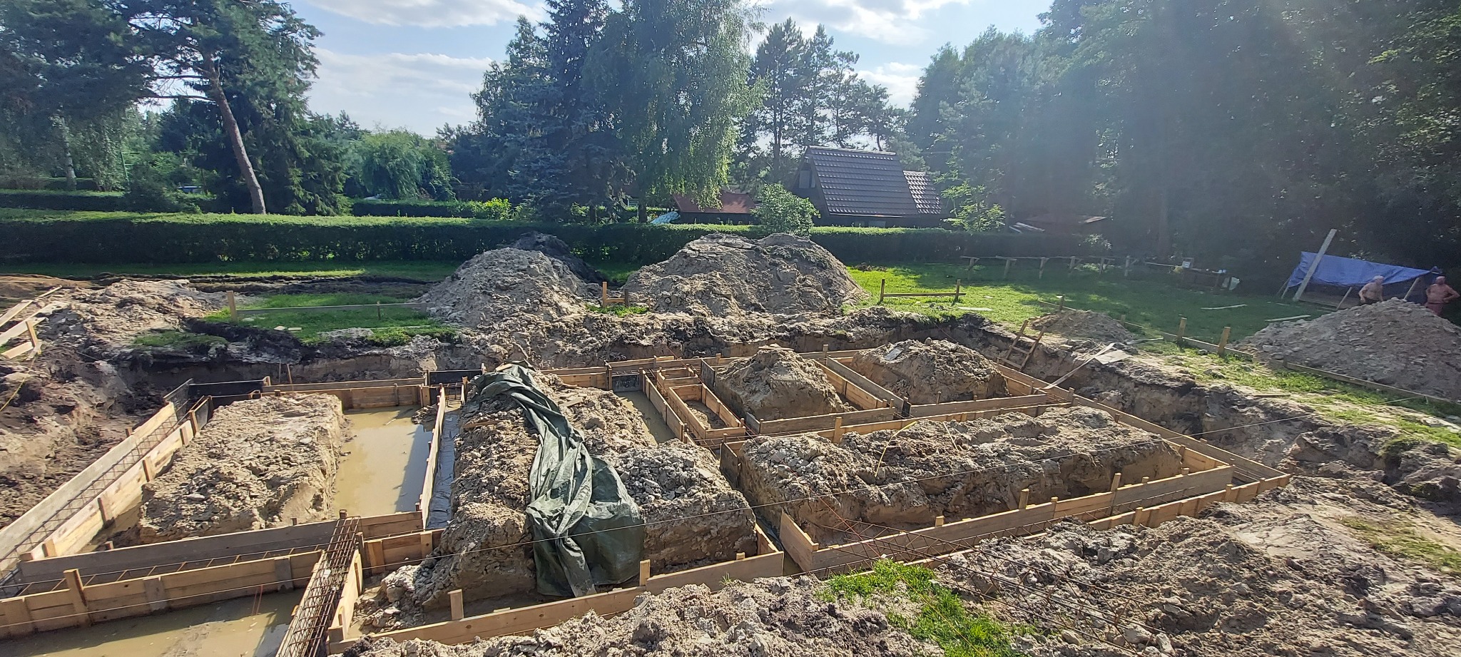 Przygotowanie terenu pod fundamenty: drewniane szalunki wypełnione ziemią i wodą, widoczne zbrojenia, w tle sterta ziemi, zieleń drzew i częściowo widoczny dach domu.