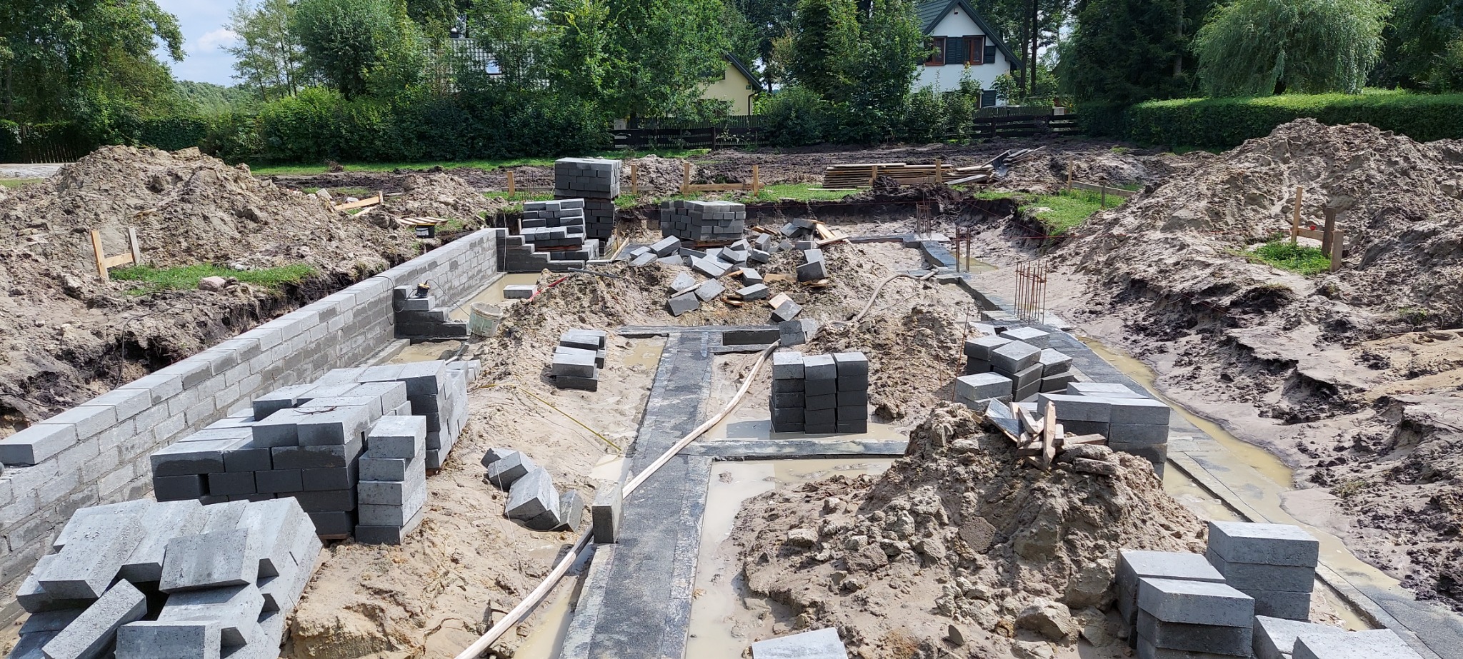 Wykop pod fundamenty z częściowo wzniesionymi ścianami z bloczków betonowych, stosy bloczków obok wykopu, teren budowy z usypaną ziemią i widoczną w oddali zabudową jednorodzinną.