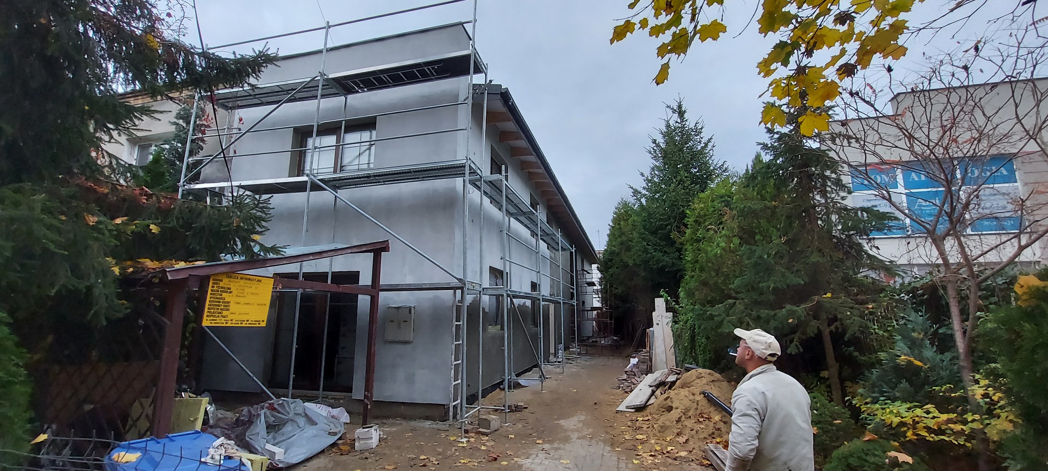 Budynek w trakcie ocieplania elewacji, otoczony rusztowaniami, z widocznym pracownikiem w oddali oraz żółtą tablicą informacyjną i jesiennym krajobrazem w tle.