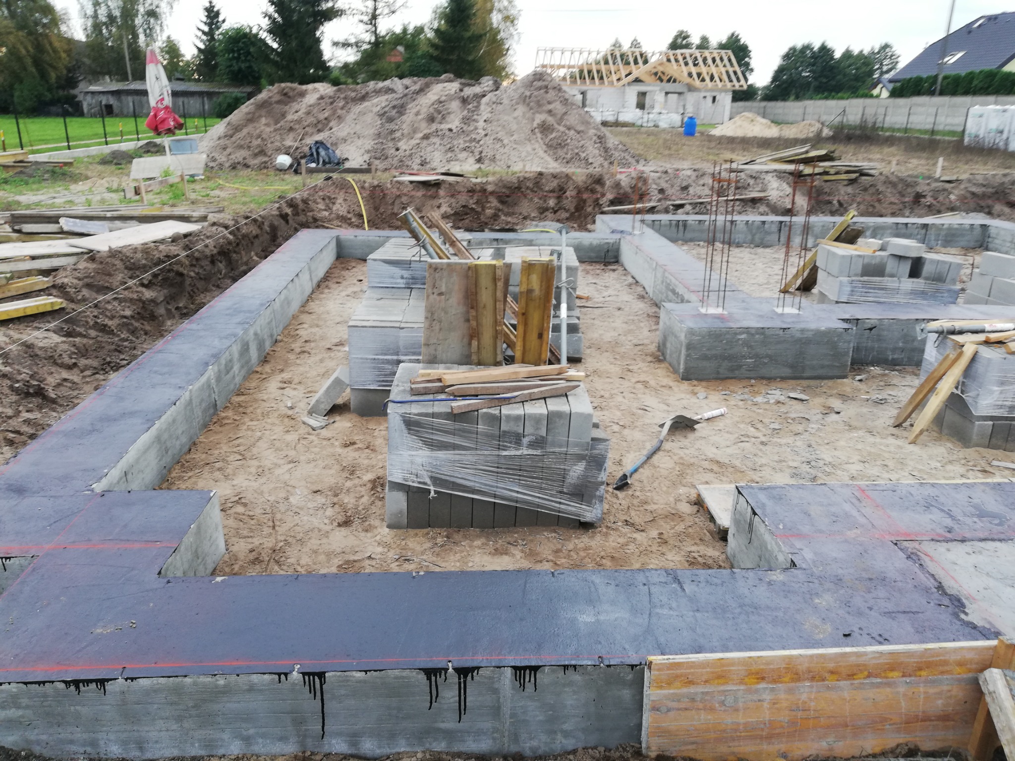Wylane fundamenty domu jednorodzinnego z widoczną izolacją przeciwwilgociową, bloczki betonowe na palecie, w tle plac budowy z hałdą ziemi i częściowo wybudowanym domem w stanie surowym otwartym.