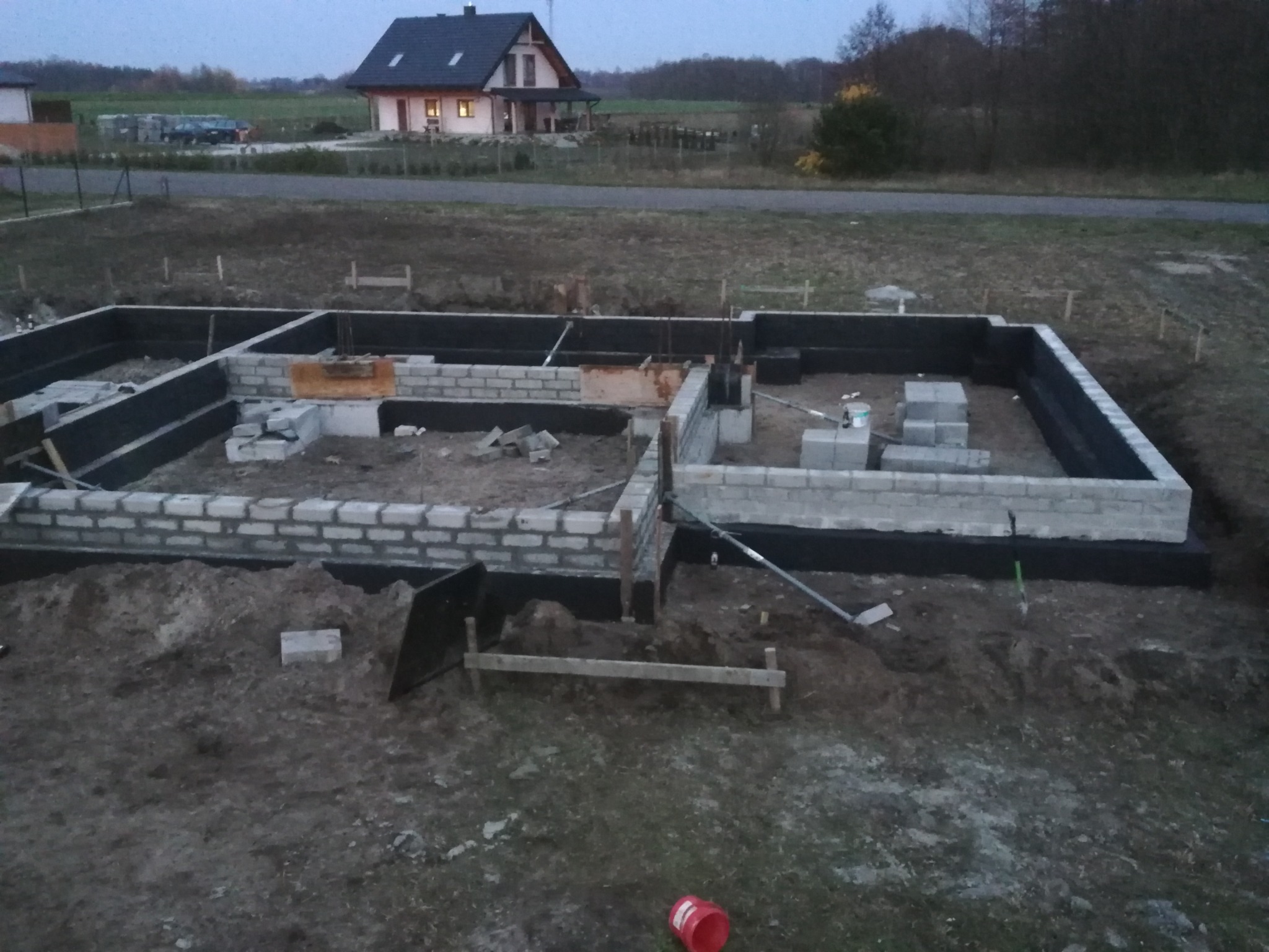 Wykopane i częściowo wymurowane fundamenty pod budowę domu jednorodzinnego, zabezpieczone czarną izolacją, w tle widoczny ukończony dom z oświetlonym oknem.