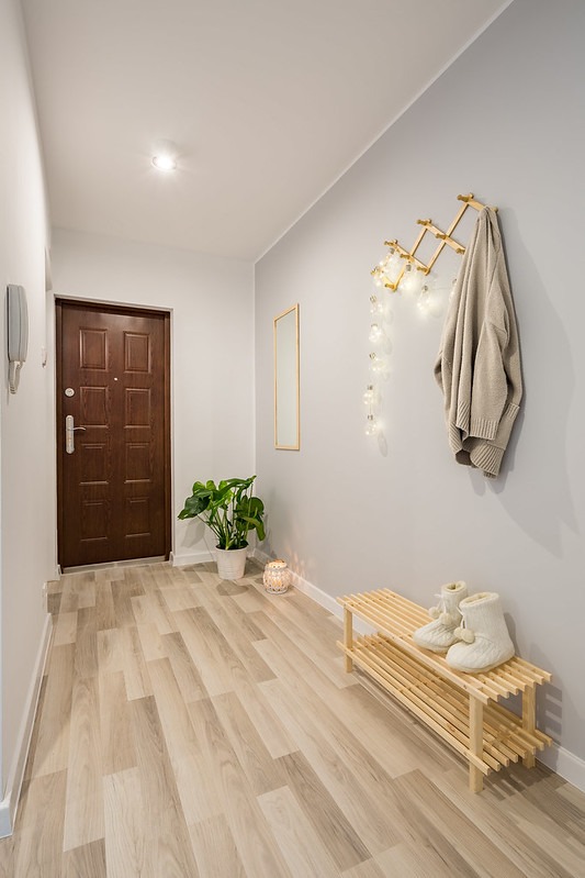 Jasny przedpokój z panelami podłogowymi imitującymi drewno, drewnianą szafką na buty i wieszakiem na ścianie, oświetlony girlandą z żarówek.