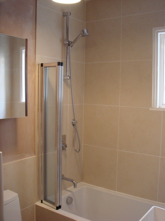 Wykończona łazienka z beżowymi płytkami, białą wanną i szklanym parawanem nawannowym, widoczny prysznic z regulowaną wysokością.