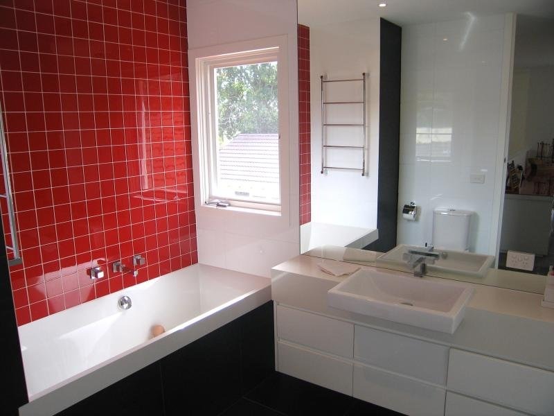 Nowoczesna łazienka z czerwoną ścianą wyłożoną kwadratowymi płytkami, białą wanną, umywalką nablatową i lustrem odbijającym toaletę oraz grzejnik drabinkowy.