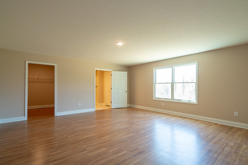 Pusty pokój z drewnopodobną podłogą, świeżo pomalowanymi ścianami w odcieniach beżu, białymi listwami przypodłogowymi, widokiem na garderobę z metalowymi półkami i otwarte drzwi do łazienki.