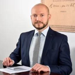 Portret mężczyzny w garniturze siedzącego za biurkiem z dokumentami i długopisem, w tle fragment starego dokumentu.