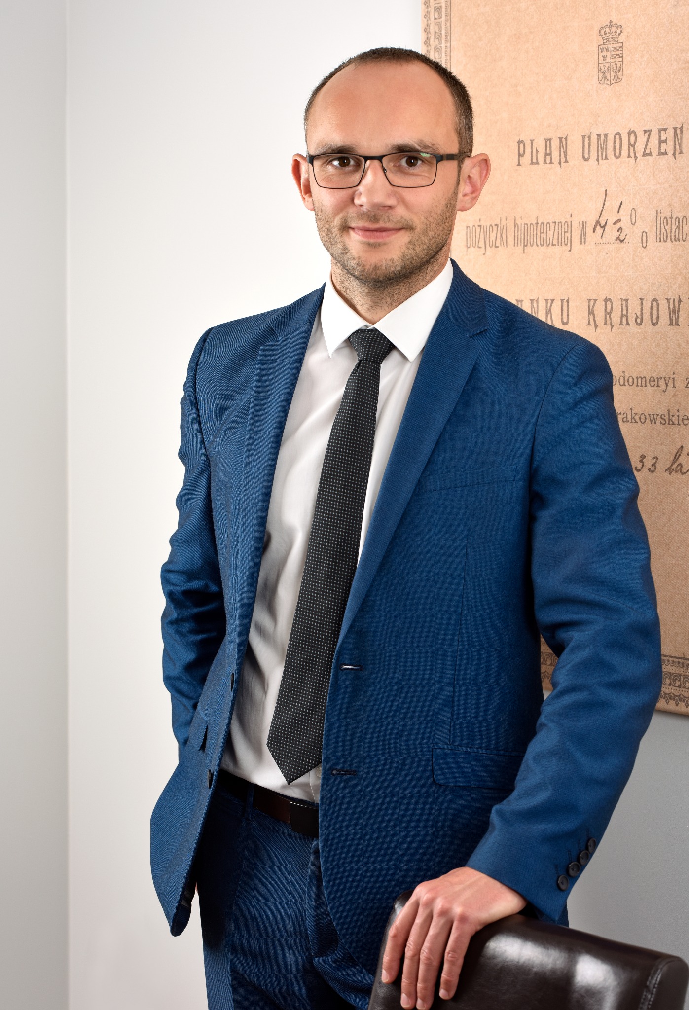Portret mężczyzny w okularach i niebieskim garniturze, opierającego się o krzesło, na tle ściany z napisem 'Plan umorzeń pożyczki hipotecznej w 4,5%'