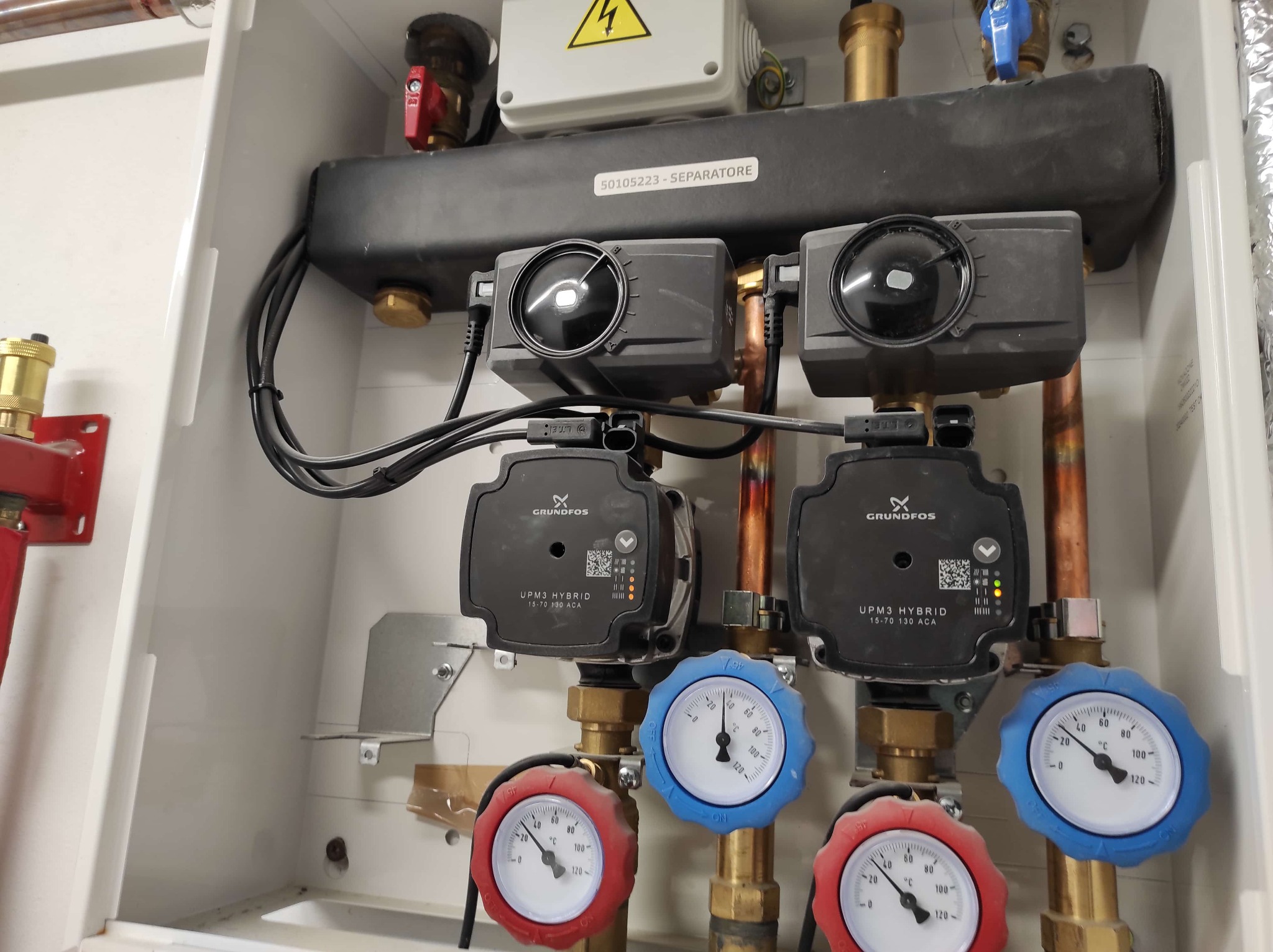Rozdzielacz hydrauliczny z dwoma pompami obiegowymi Grundfos UPM3 Hybrid, manometrami z czerwonymi i niebieskimi pokrętłami, oraz separatorem powietrza, zamontowany w białej szafce instalacyjnej.