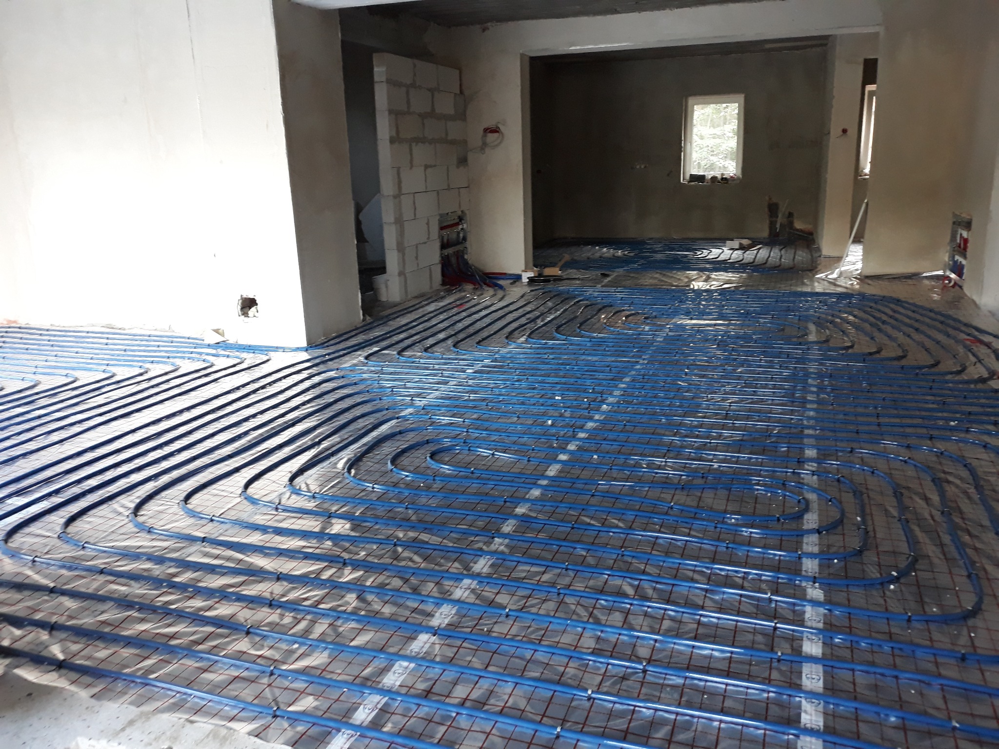 Niebieskie rury ogrzewania podłogowego ułożone w pętle na folii odblaskowej w surowym wnętrzu budynku w trakcie budowy.