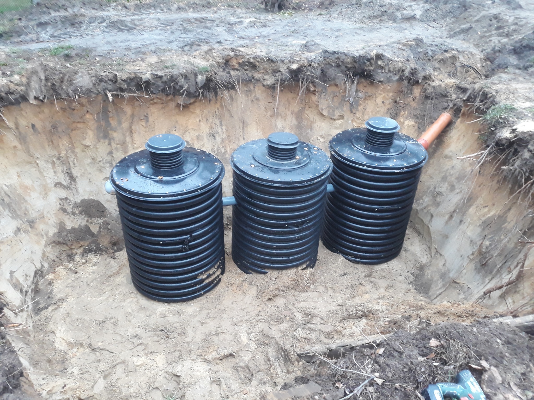 Instalacja trzech czarnych, plastikowych zbiorników bezodpływowych w wykopie ziemnym, widoczny fragment rury kanalizacyjnej i zakurzona wiertarko-wkrętarka na pierwszym planie.