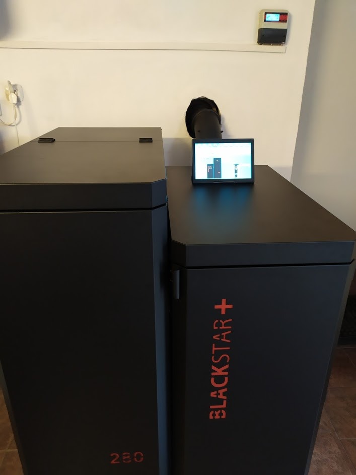 Nowoczesny, czarny piec na pellet Blackstar+ z panelem sterowania dotykowym, podłączony do komina, zamontowany w jasnym pomieszczeniu.