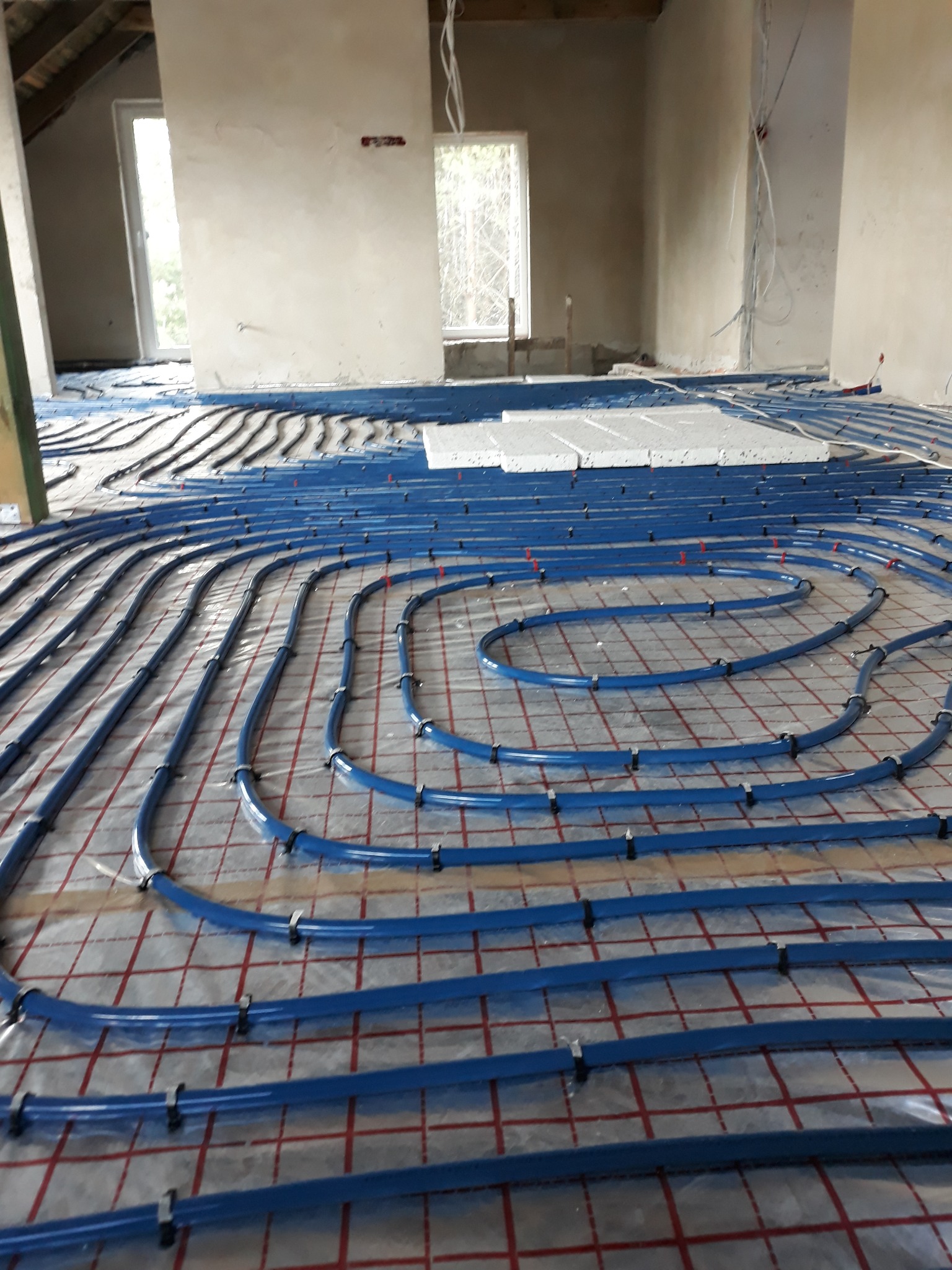 Niebieskie rury ogrzewania podłogowego ułożone w pętle na folii z siatką w surowym wnętrzu budynku, widoczne okna i nieotynkowane ściany.