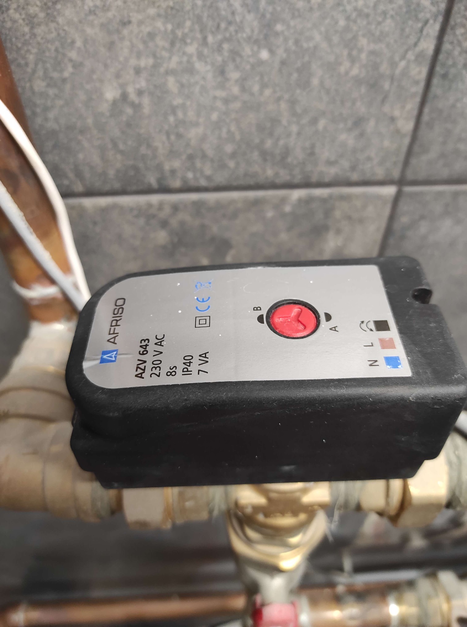 Zawór AFRISO AZV 643 z czerwonym pokrętłem, zamontowany na miedzianej instalacji grzewczej, na tle szarej ściany.