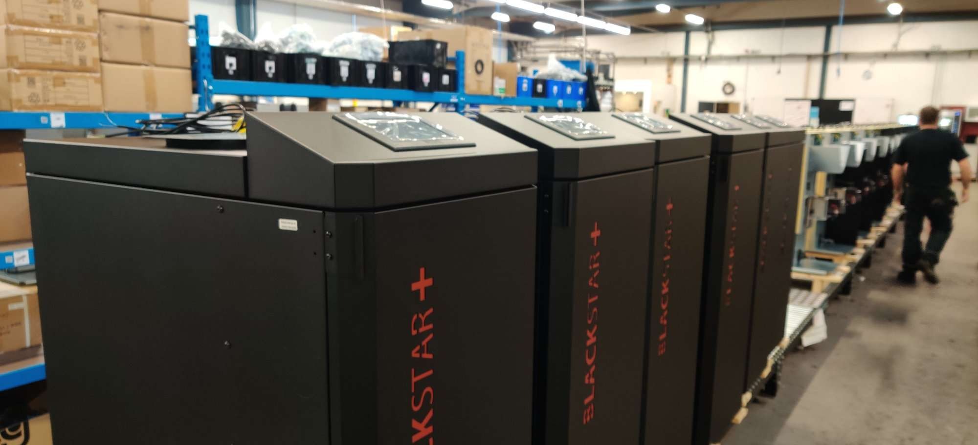 Rząd czarnych kotłów grzewczych BLACKSTAR+ z dotykowymi panelami sterowania, ustawionych w magazynie lub hali produkcyjnej, widoczny pracownik w tle.