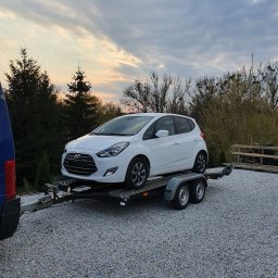 BST Aneta Wojnarowska - Biały samochód osobowy Hyundai i20 na dwuosiowej przyczepie lawetowej z widocznym fragmentem niebieskiego samochodu transportowego i żwirowym podłożem