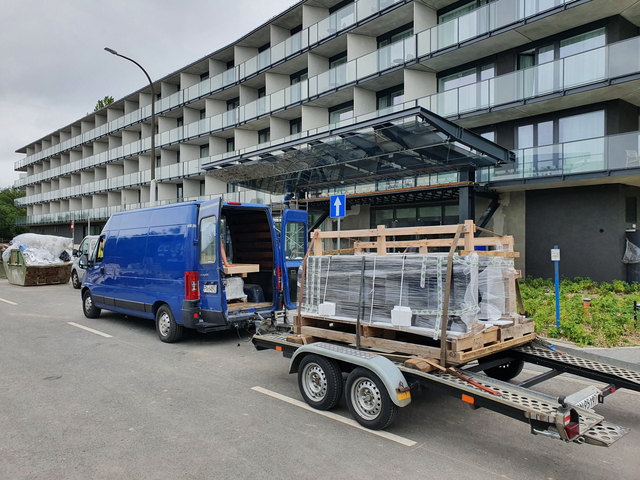 Niebieski bus z otwartymi tylnymi drzwiami i przyczepą z ładunkiem okien na palecie, zaparkowany przed nowoczesnym budynkiem z balkonami.