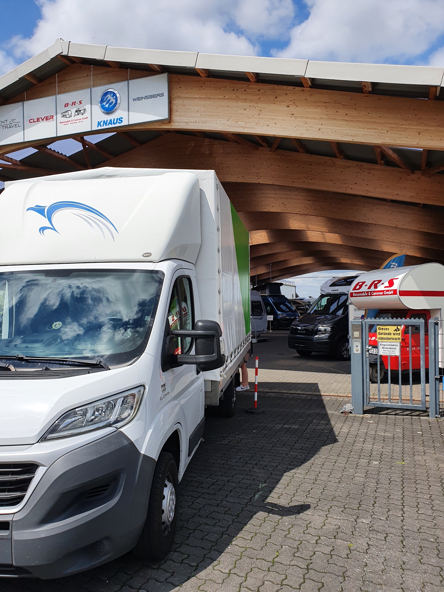 Biały samochód dostawczy z plandeką i logo orła na tle hali z drewnianą konstrukcją dachu i szyldami reklamowymi firm z branży caravaningowej.