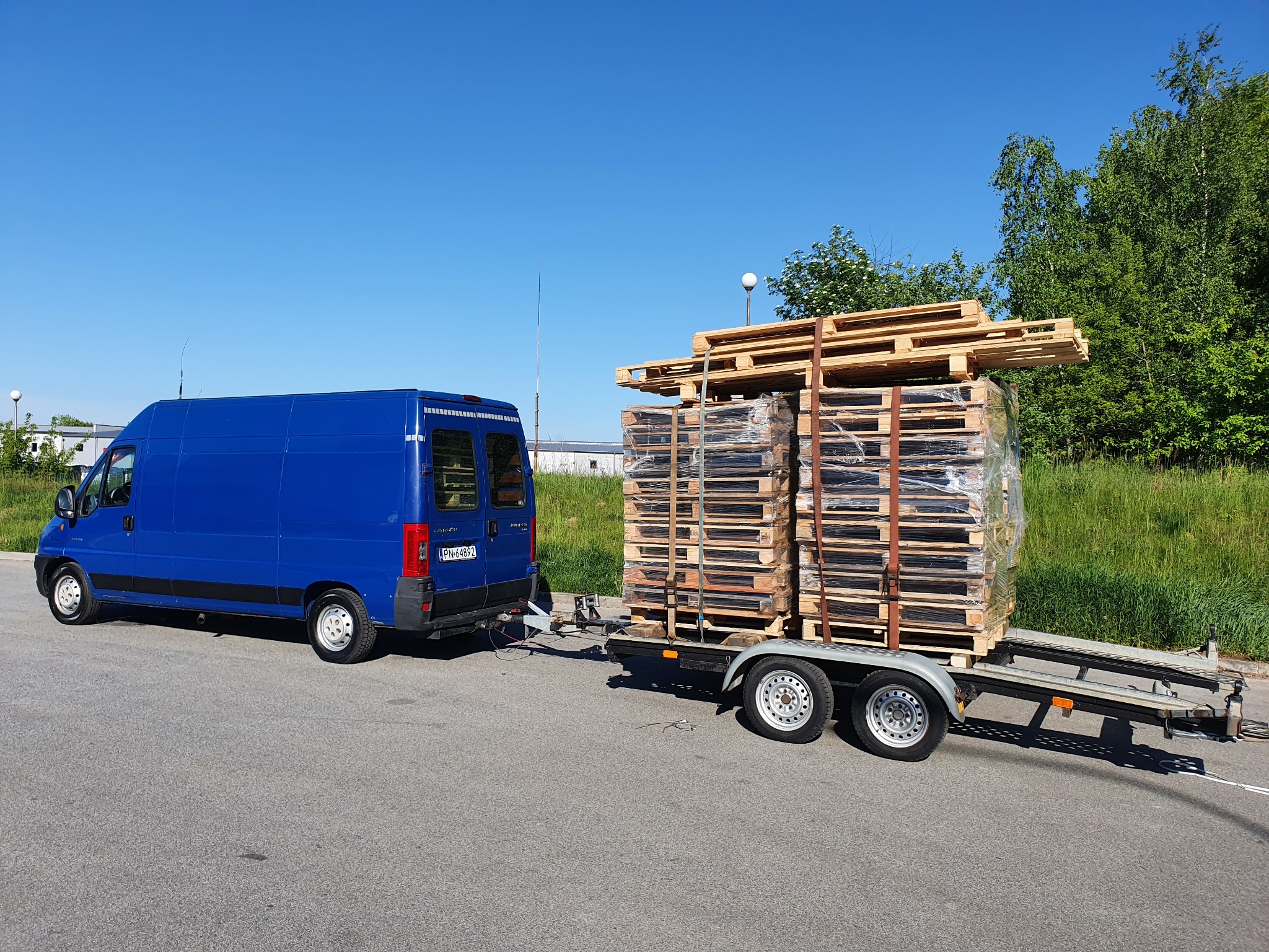 Niebieski bus z przyczepą przewożący palety, zabezpieczone folią stretch i pasami transportowymi, na tle zieleni i błękitnego nieba.
