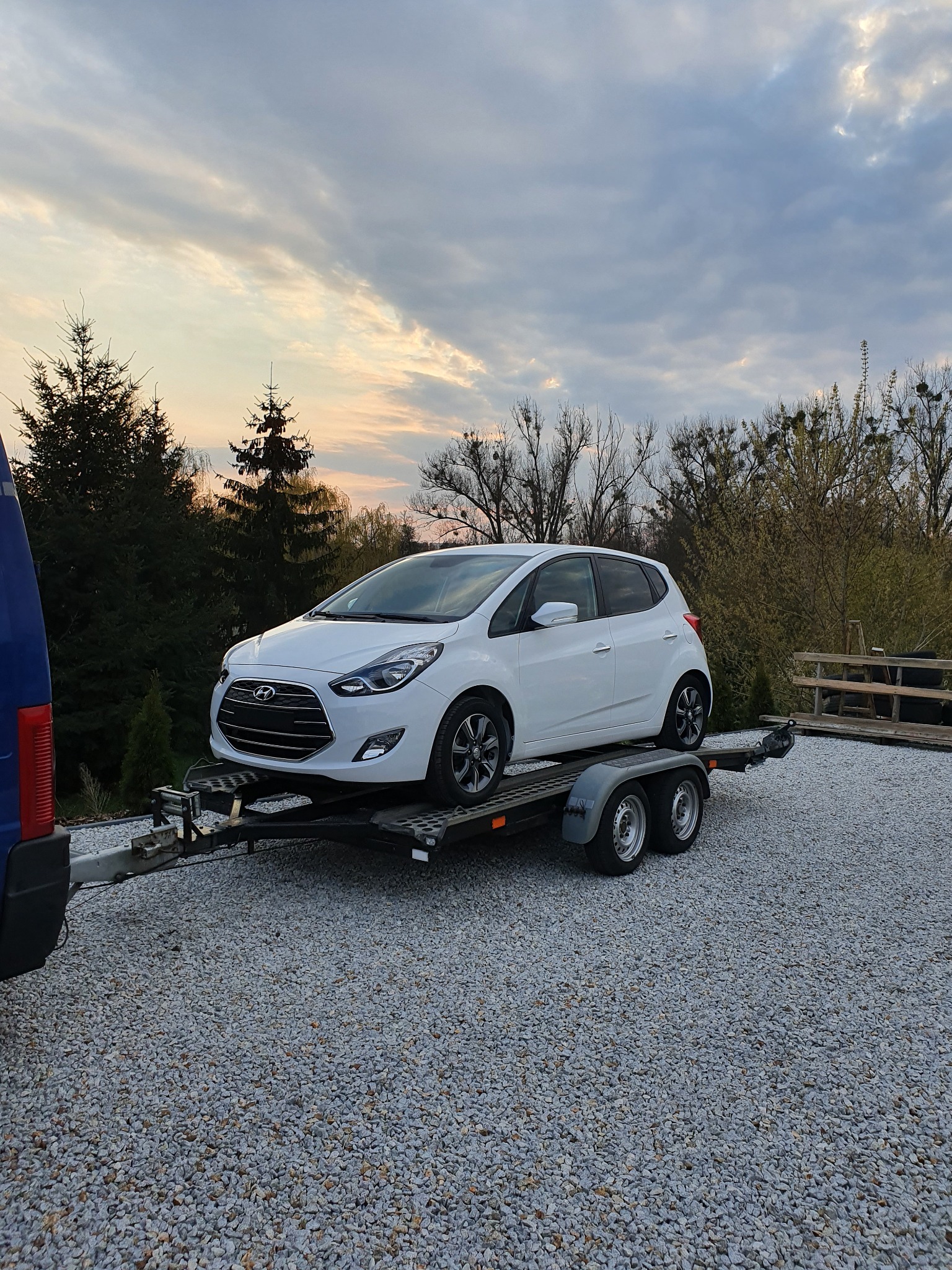 Biały samochód osobowy Hyundai i20 na dwuosiowej przyczepie lawetowej z widocznym fragmentem niebieskiego samochodu transportowego i żwirowym podłożem