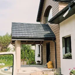 Nowoczesny dom z ciemną dachówką i drewnianą elewacją, widok z boku z elementami ogrodu i samochodu dostawczego w kadrze. Widoczne detale wykończenia.