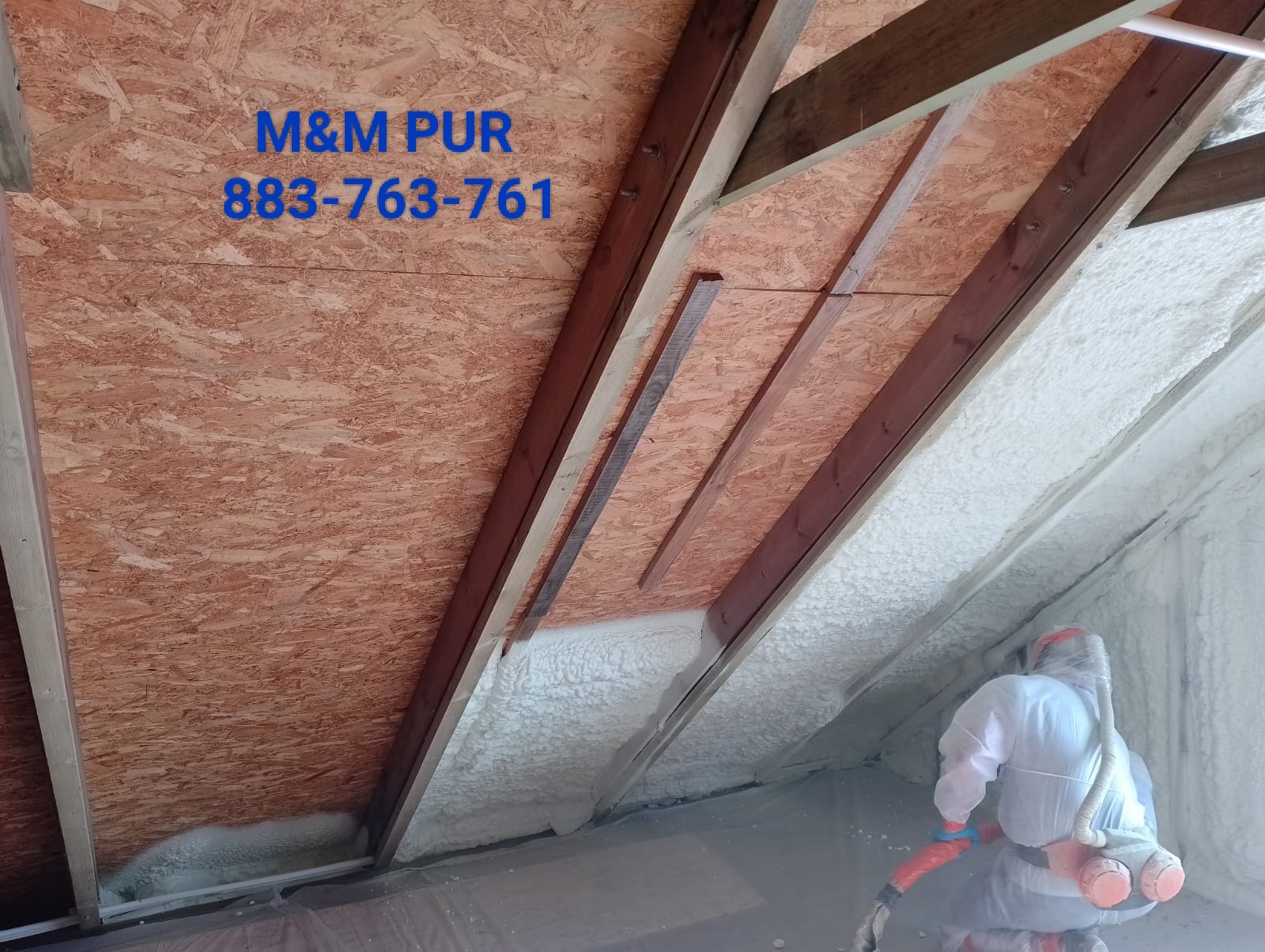 Poddasze ocieplane pianką natryskową, widoczny pracownik w kombinezonie ochronnym aplikujący izolację między krokwiami. Na ścianie widoczny napis 'M&M PUR 883-763-761'.