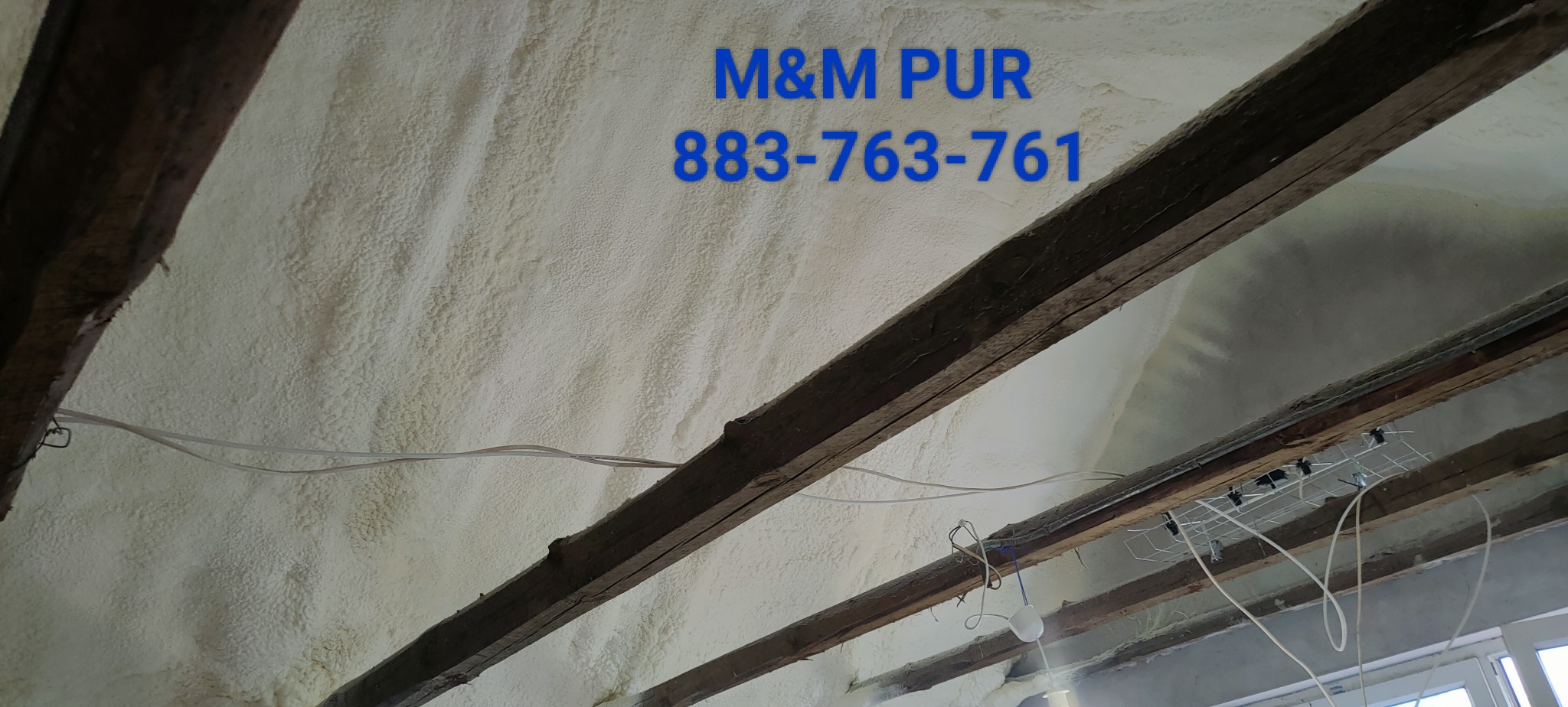 Poddasze ocieplone pianką, widoczne drewniane belki stropowe i instalacja elektryczna, na ścianie nadrukowane logo 'M&M PUR' wraz z numerem telefonu.