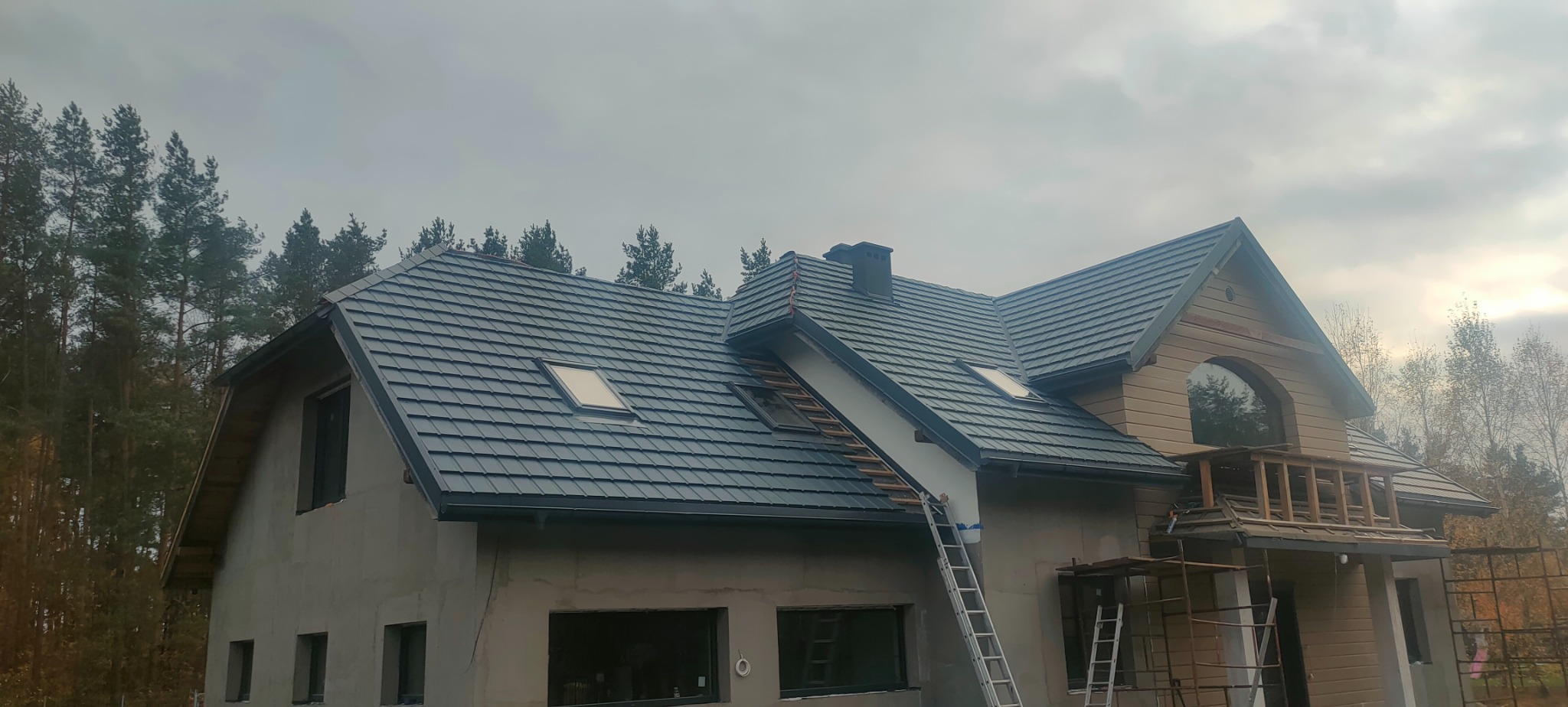 Dom w trakcie budowy z nowym dachem z dachówki ceramicznej w kolorze grafitowym, widoczne okna dachowe i drabina oparta o ścianę, elewacja w stanie surowym.