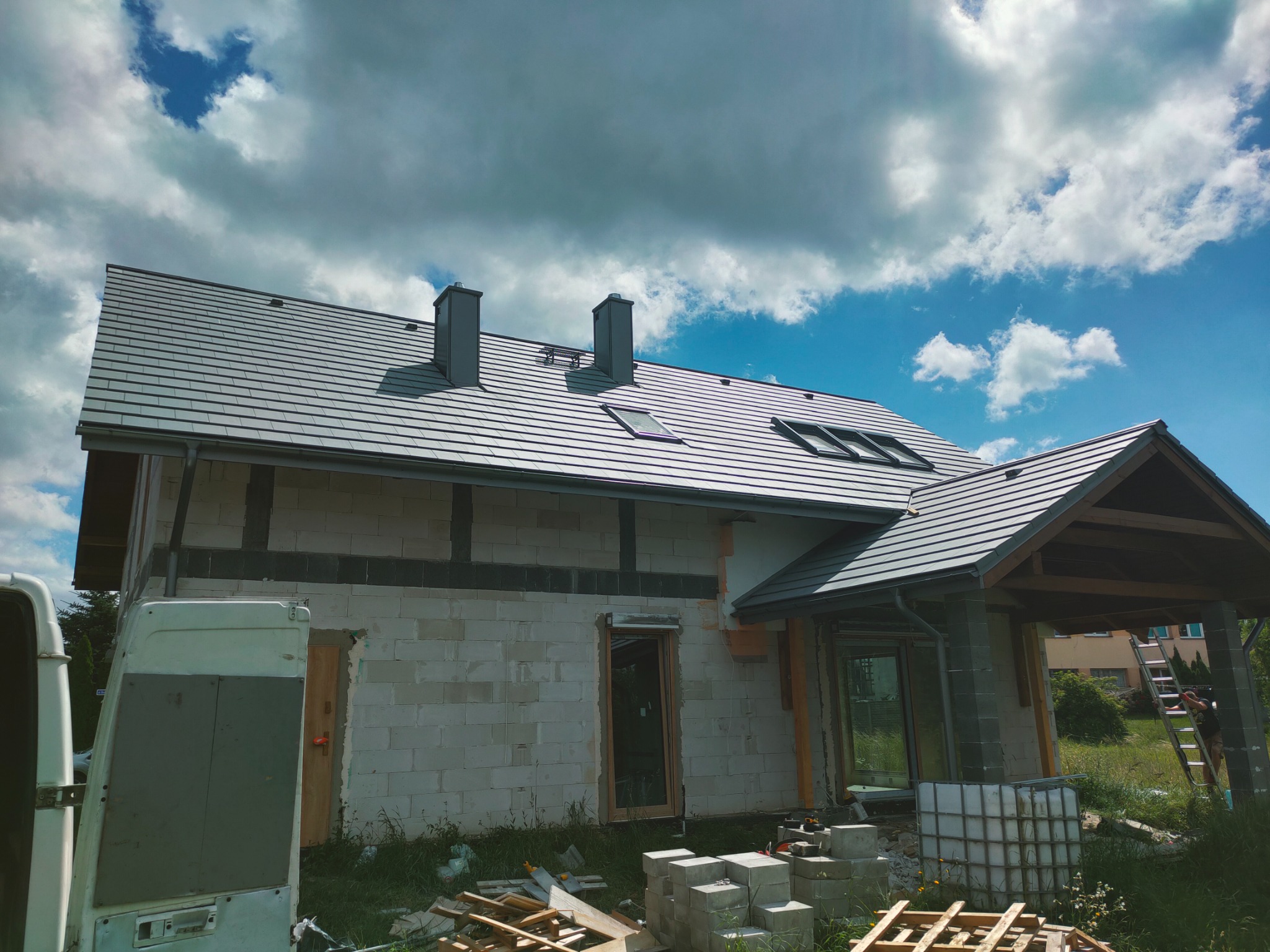 Nowy dom w trakcie budowy z szarą dachówką, widoczne okna dachowe i kominy, ściany z białego bloczka, częściowo ocieplone, na pierwszym planie materiały budowlane i samochód dostawczy.