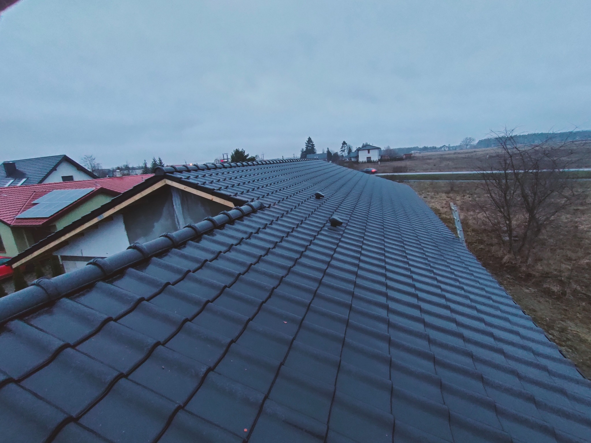 Nowy, czarny dach z dachówki ceramicznej, widok z góry na tle pochmurnego nieba i krajobrazu podmiejskiego.