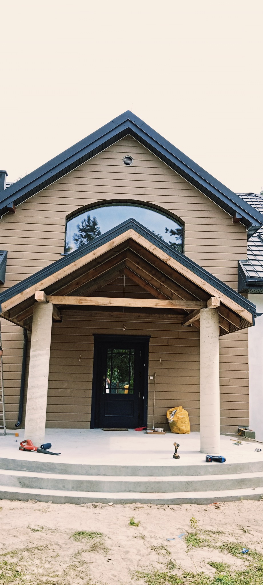 Dom z brązową elewacją i czarną stolarką okienną. Widoczny zadaszony ganek z betonowymi kolumnami, narzędzia budowlane i materiały na schodach.