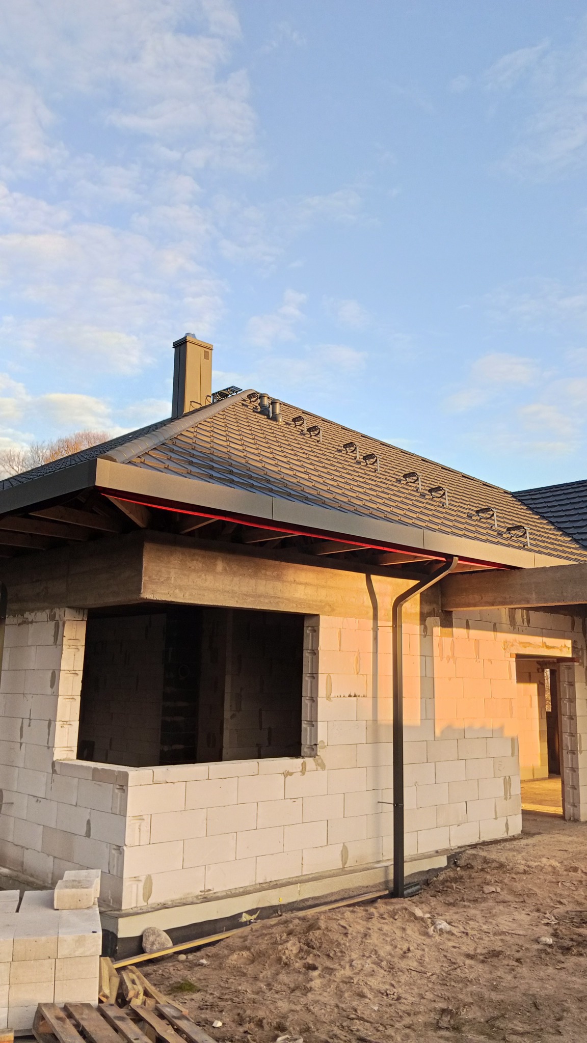 Surowy stan domu z szarą dachówką, czarną rynną i kominem na tle błękitnego nieba. Widoczne bloczki betonowe i niezabudowane otwory okienne.