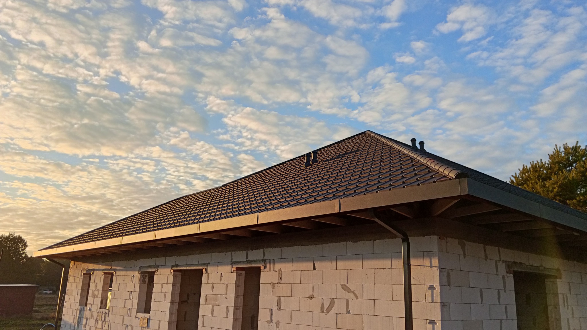 Nowy, brązowy dach z blachodachówki modułowej na surowym budynku w Bydgoszczy, widok z dołu na tle błękitnego nieba z obłokami. Widoczne kominy wentylacyjne.