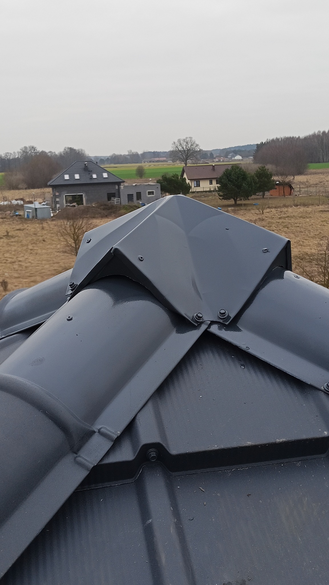 Szary dach z blachy trapezowej z widocznym systemem mocowania na tle krajobrazu wiejskiego i zabudowań. Widok z góry, pochmurny dzień.