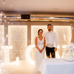 Para młoda stoi za tortem weselnym na tle dekoracji świetlnych z napisem LOVE i fontannami iskier.