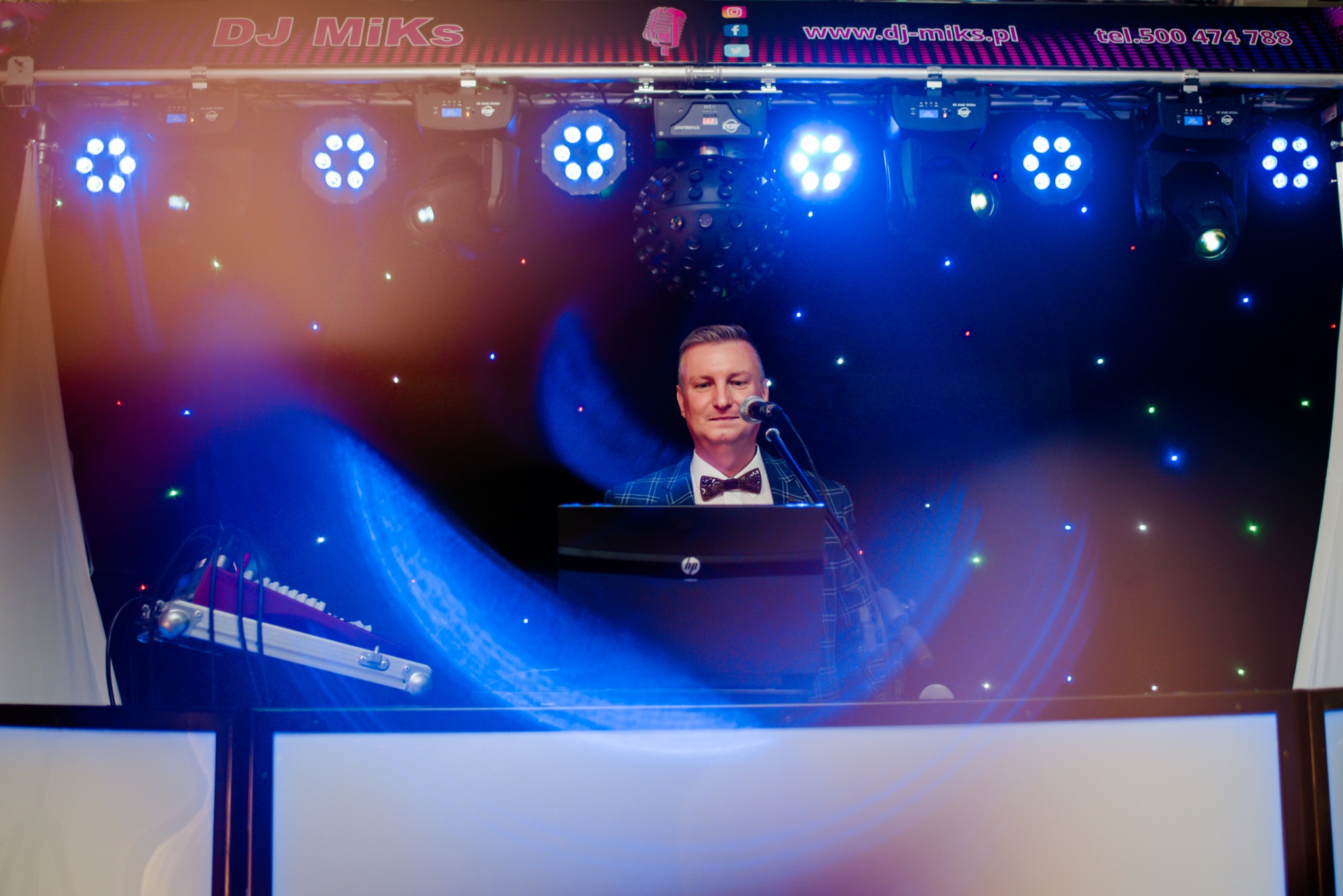 DJ w marynarce w kratę i muszce, stojący za konsoletą z laptopem, oświetlony niebieskimi światłami scenicznymi na tle czarnej ściany z gwiazdkami, nad nim baner z nazwą DJ Miks i danymi kontaktowymi.