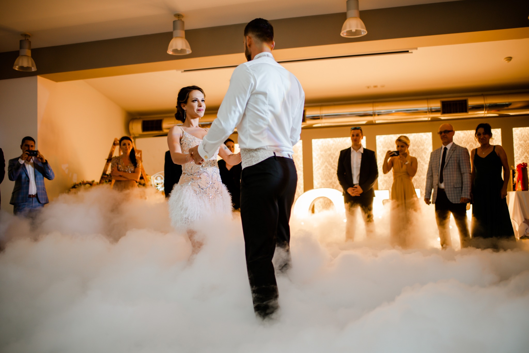 Para młoda podczas pierwszego tańca w chmurach, goście obserwują i nagrywają, dekoracje ślubne z podświetlanymi literami w tle.