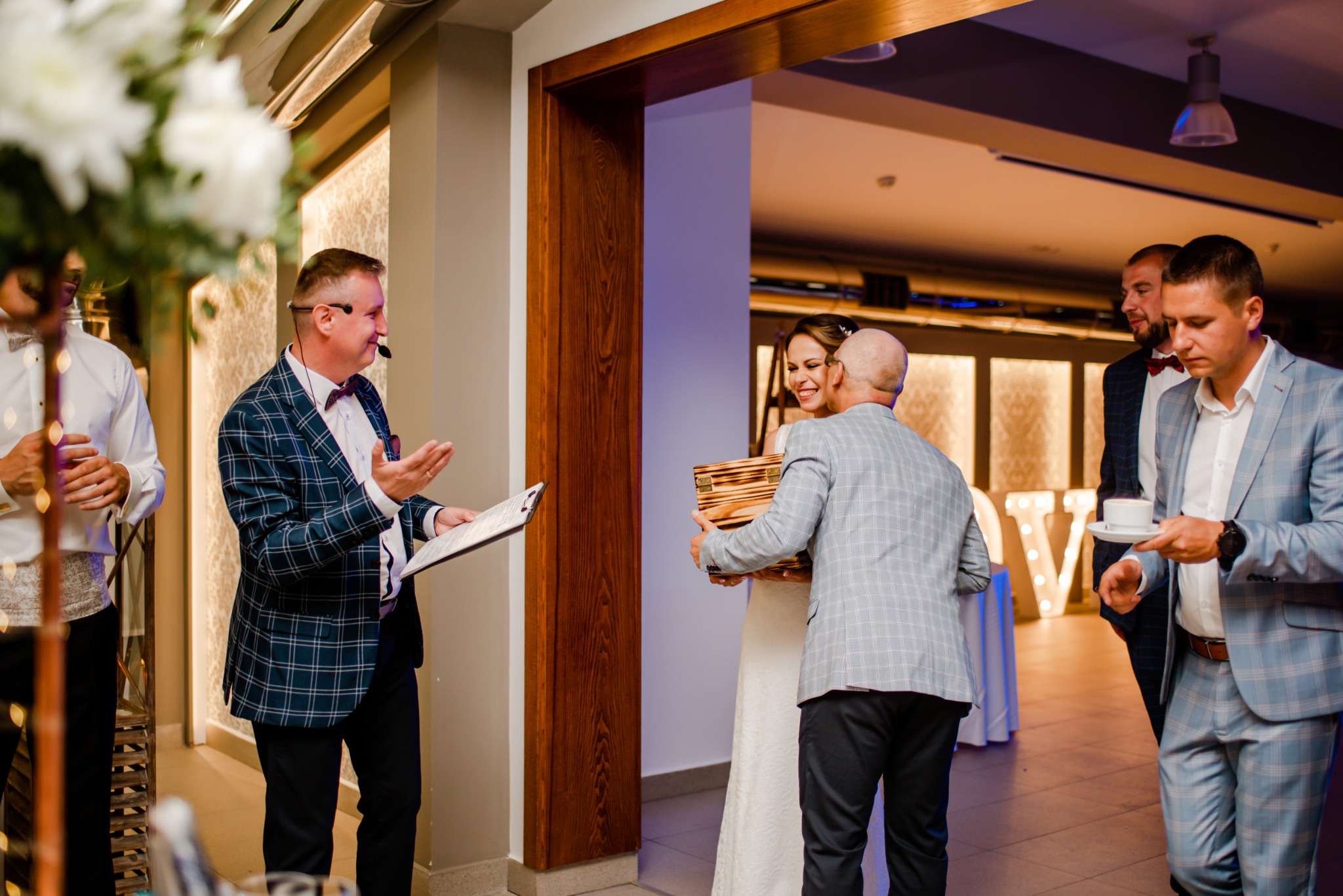 Prezenter w kraciastej marynarce z mikrofonem na głowie prowadzi ceremonię wręczania prezentu młodej parze, w tle goście weselni i podświetlane litery układające się w napis.