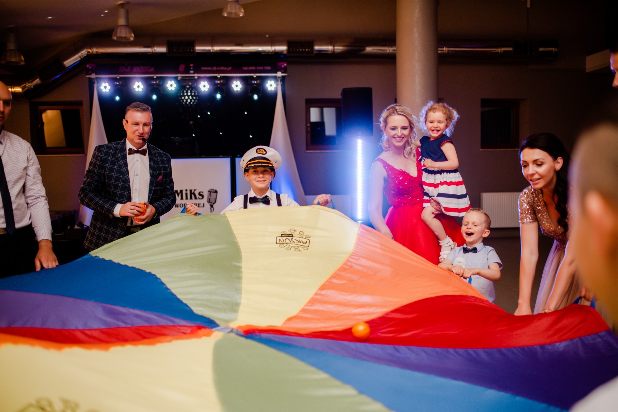 Animatorzy prowadzą zabawy z dziećmi przy użyciu kolorowej chusty animacyjnej w sali z dekoracjami świetlnymi.