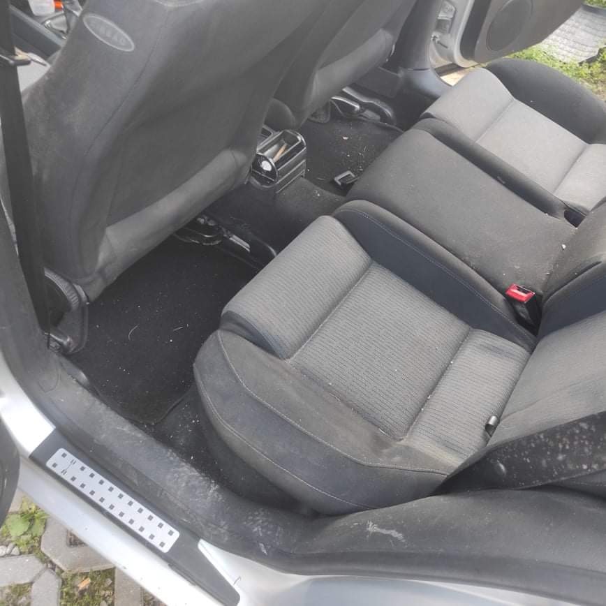 Wnętrze samochodu z widocznymi, szarymi fotelami tylnymi, dywanikami i progami, zanieczyszczone kurzem i drobnymi zabrudzeniami przed czyszczeniem.