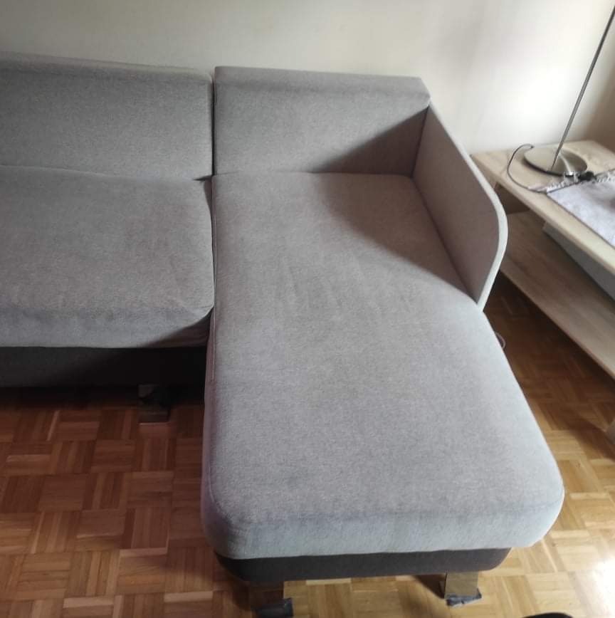 Jasnoszara, narożna sofa z tapicerką, widoczna z boku, stojąca na parkiecie. Obok stolik z lampą.