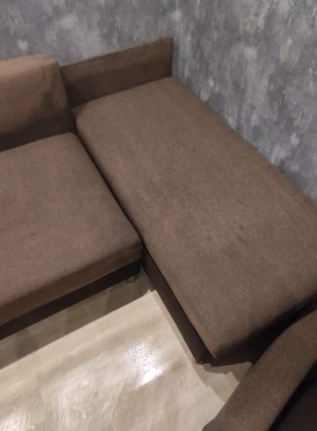 Część brązowej, modułowej kanapy z widocznymi śladami użytkowania na siedzisku i oparciu, stojąca na jasnej podłodze przy szarej ścianie.