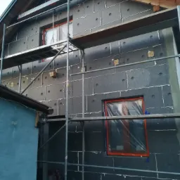 Dom w trakcie ocieplania: grafitowy styropian na elewacji, rusztowania, okna zabezpieczone folią, widoczne worki z materiałami budowlanymi.