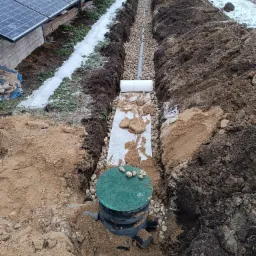 Wykopany rów z ułożonymi rurami drenarskimi i studzienką rewizyjną z zieloną pokrywą, zasypany częściowo żwirem; w tle panele solarne i pracownik przy dalszej części instalacji.