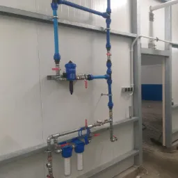 Niebiesko-srebrna instalacja wodna z filtrami i zaworami, zamontowana na białej ścianie z metalowymi profilami, widok z boku na fragment pomieszczenia z otwartymi drzwiami.