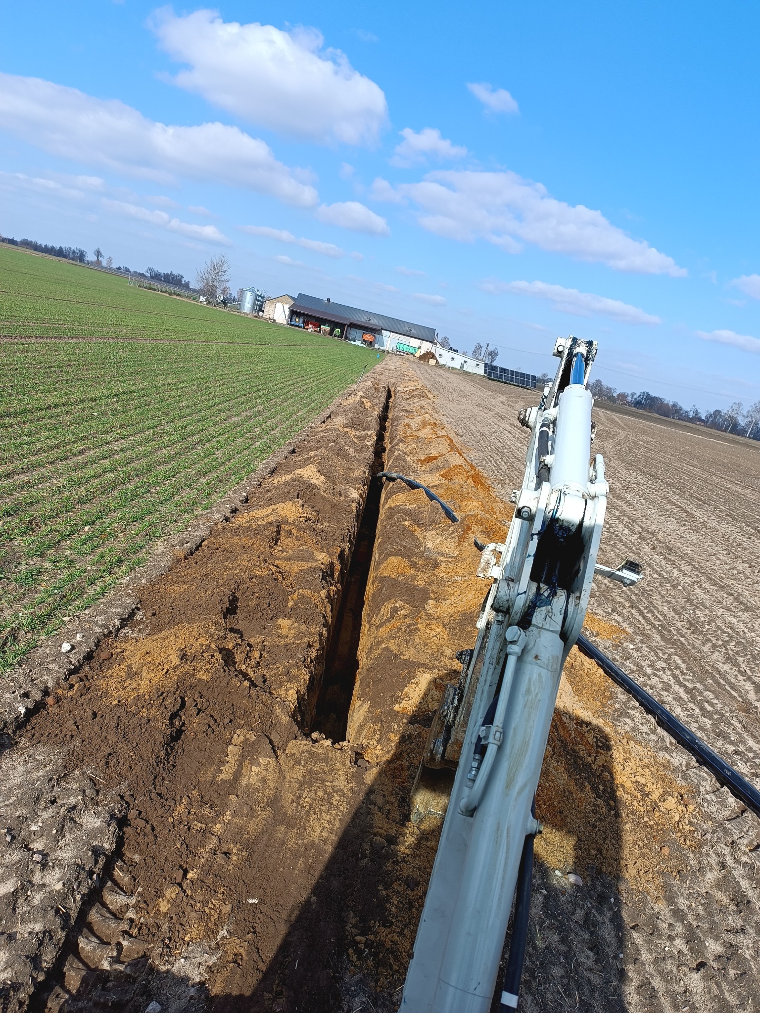 Wąski wykop w polu uprawnym, wykonany koparką widoczną z perspektywy operatora, błękitne niebo z chmurami w tle.